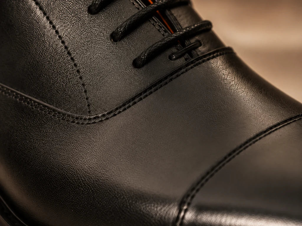 Close-up of a black leather shoe upper showing fine grain, black laces and stitched panel edges.