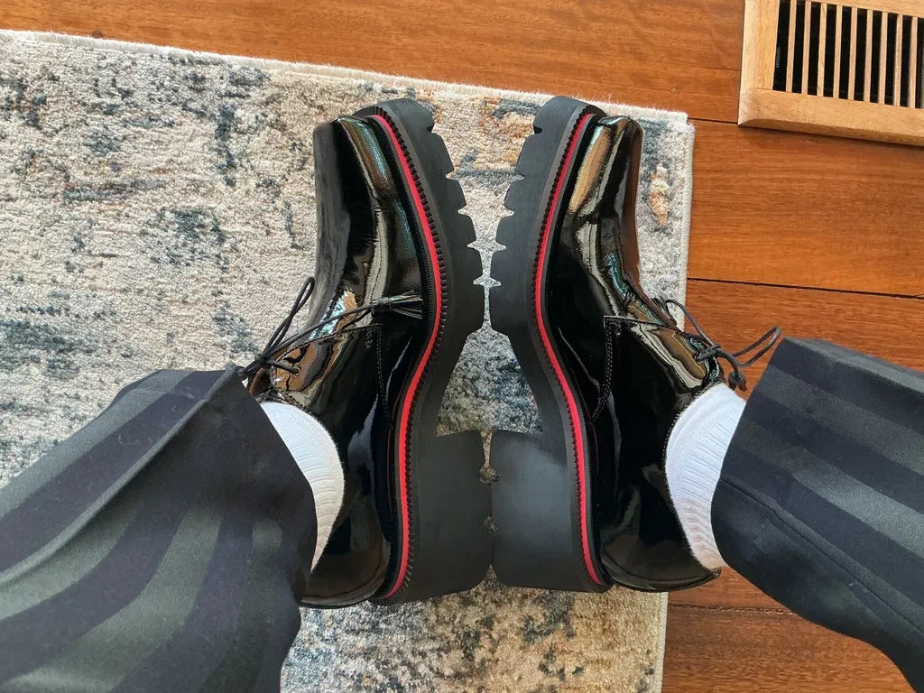 Side view of glossy black lace-up shoes with chunky lug soles and red trim, worn with striped trousers.