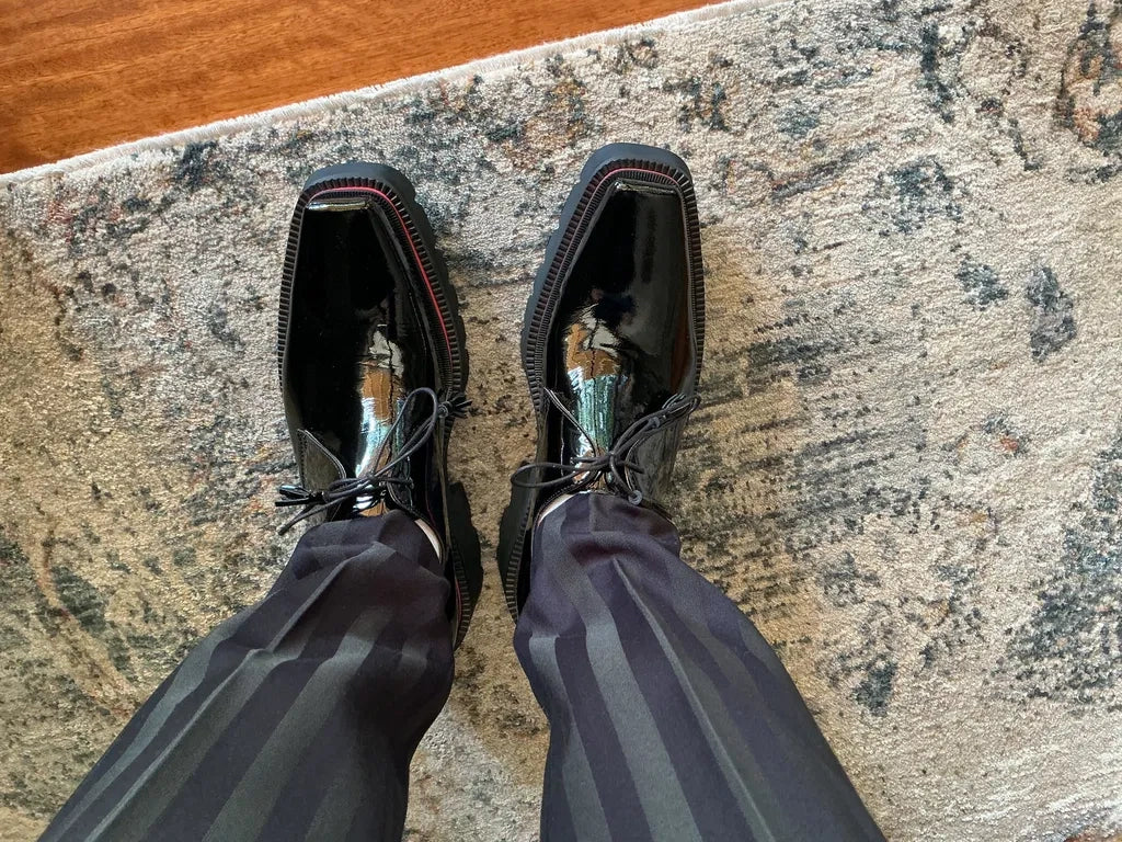 Top view of glossy black square-toe lace-up shoes worn with dark striped trousers on a patterned rug.