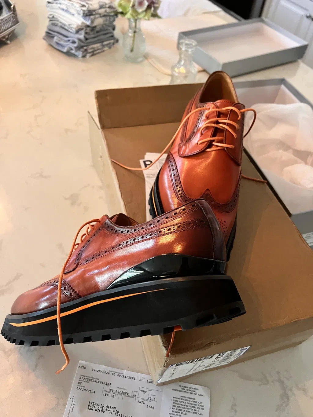 Brown leather wingtip brogues on a shipping box, showing chunky black lug soles with orange trim.