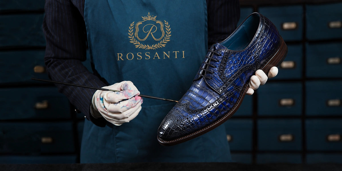 Workshop scene of a gloved artisan holding a blue wingtip brogue shoe and a fine brush, wearing a Rossanti apron.