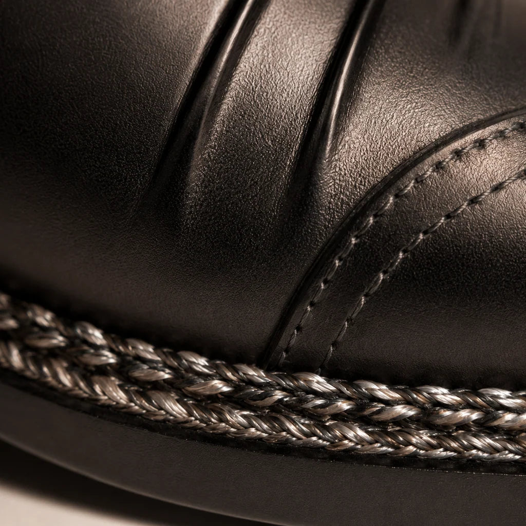 Close-up of a black leather shoe showing braided stitching along the sole edge and a stitched upper seam.