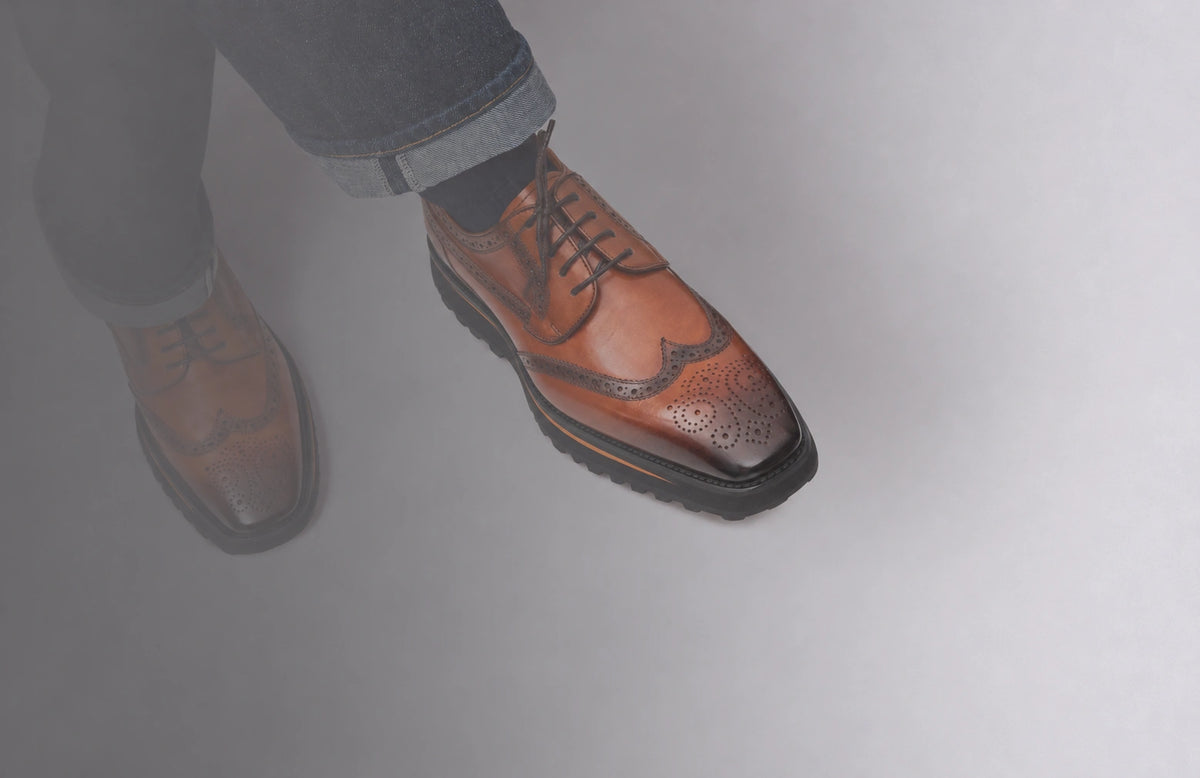 Cognac wingtip brogue shoes with black lug soles, worn with cuffed dark jeans against a softly shaded grey background.